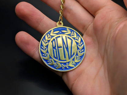 Hand holding a circular Benz enamel badge keychain to demonstrate the premium metallic weight and detailed 3D gold finish.