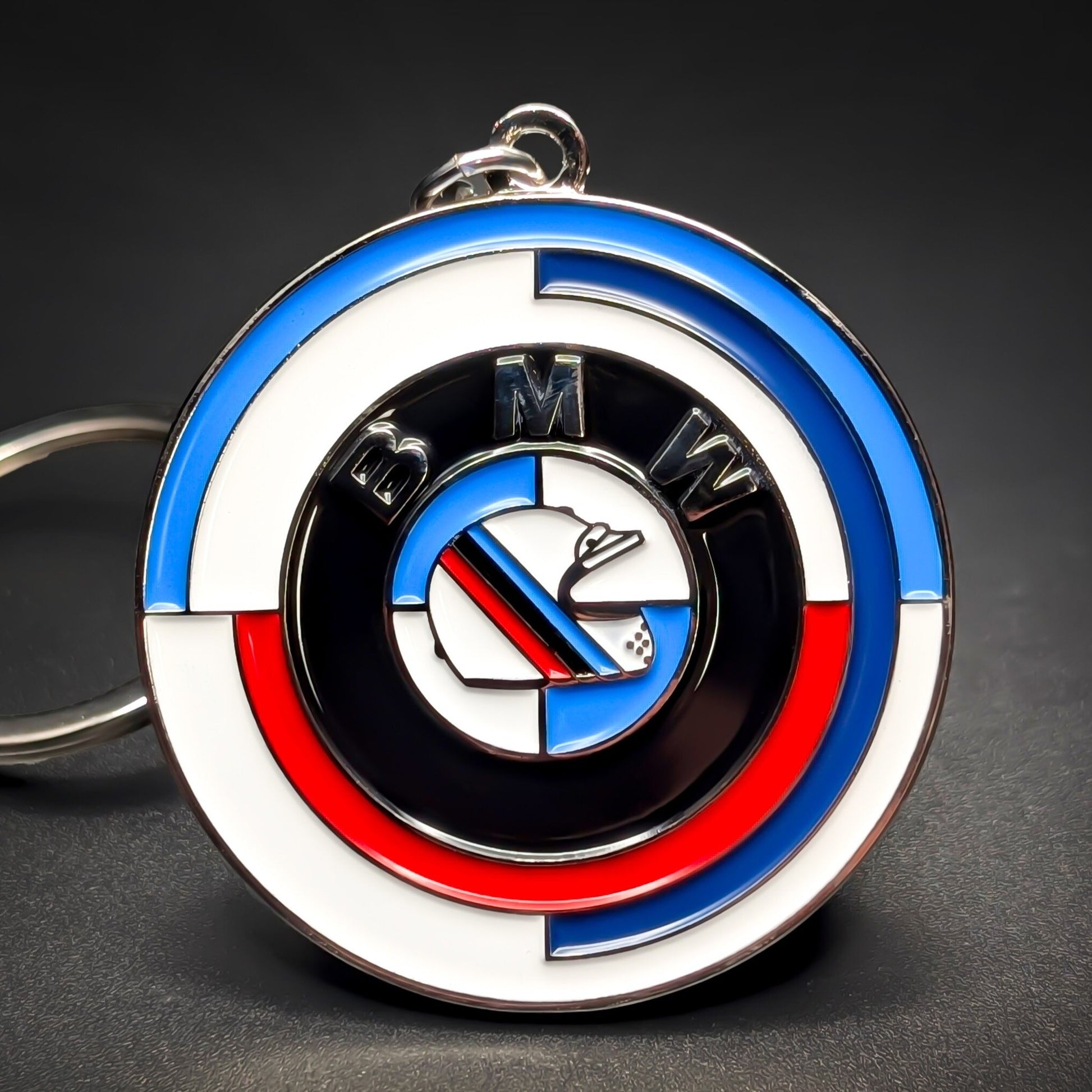 Double-sided round metal BMW badge keychain, chrome rim with blue, white, and red arced stripes, a black ring lettered BMW, and a center roundel crossed by an M-stripe band, chain at the edge, dark background.