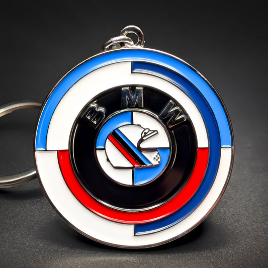 Double-sided round metal BMW badge keychain, chrome rim with blue, white, and red arced stripes, a black ring lettered BMW, and a center roundel crossed by an M-stripe band, chain at the edge, dark background.