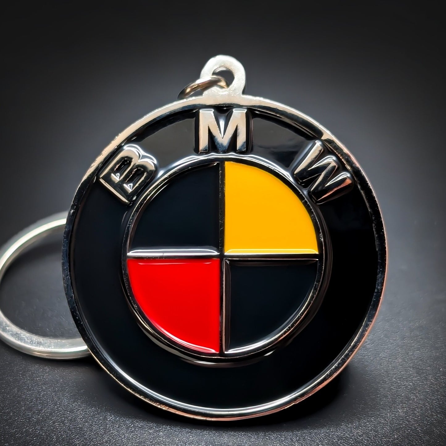 Double-sided metal BMW badge keychain with a black ring reading ‘BMW’ in silver lettering, encircling a roundel in black, gold, and red quadrants, chrome split ring, on a dark surface.