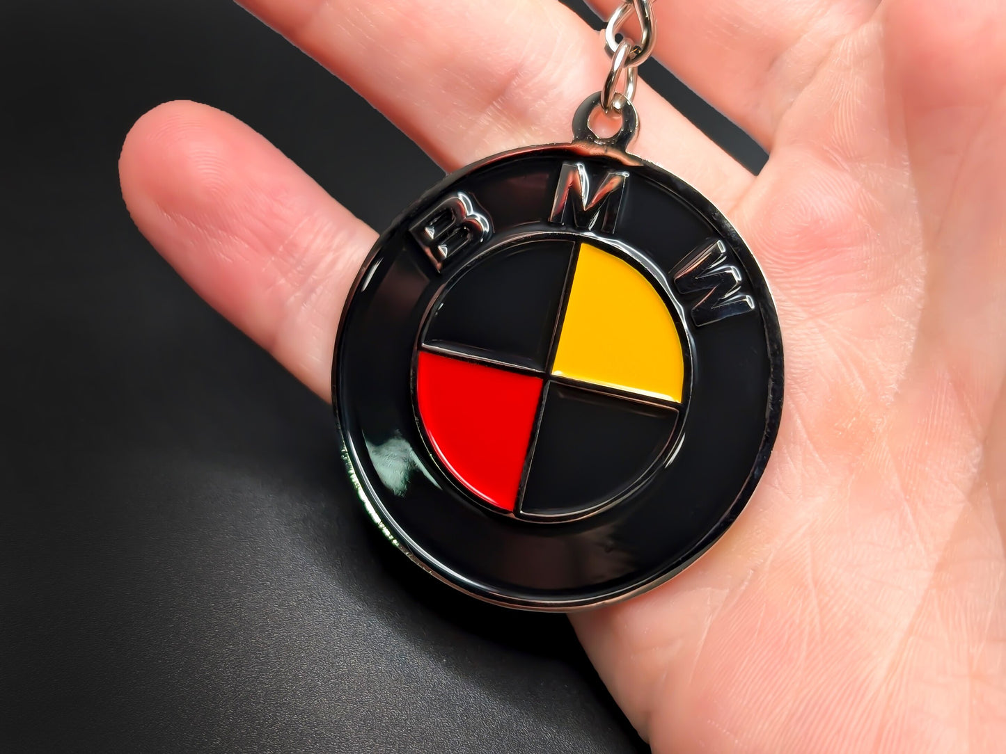 Hand holding a circular BMW enamel badge keychain to demonstrate the premium metallic weight and vibrant 3D raised lettering.