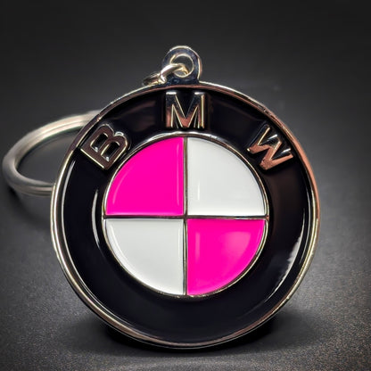 Double-sided metal BMW badge keychain with a black ring reading ‘BMW’ in silver lettering, encircling a roundel in alternating pink and white quadrants, chrome split ring, on a dark surface.
