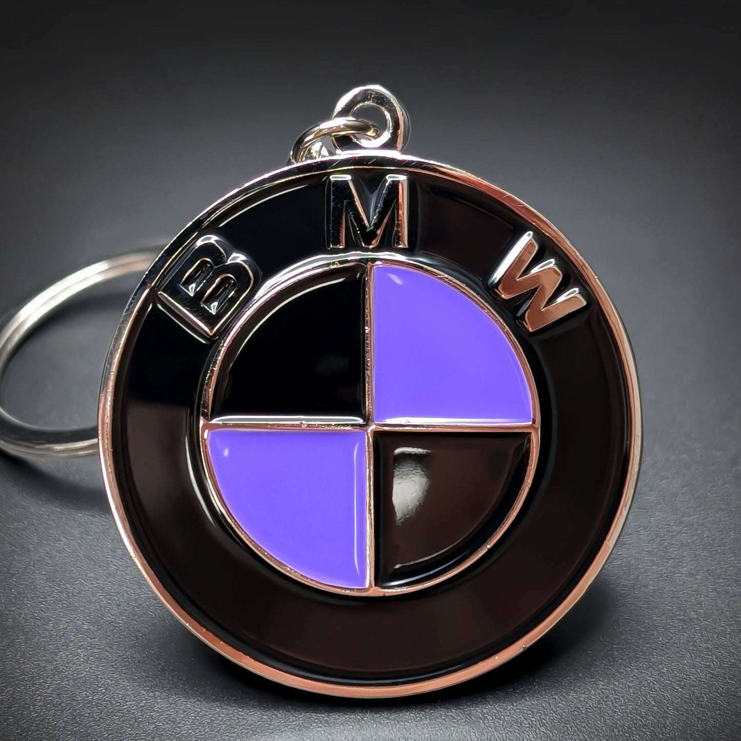 Double-sided metal BMW badge keychain with a black ring reading ‘BMW’ in silver lettering, encircling a roundel in alternating black and purple quadrants, chrome split ring, on a dark surface.