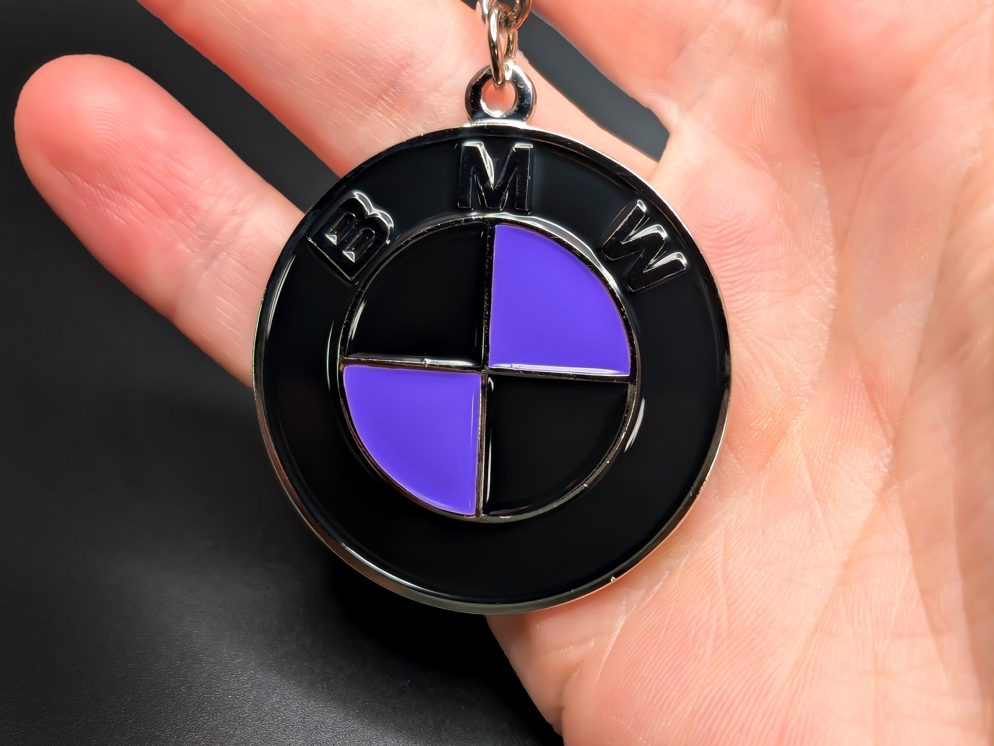 Hand holding a circular purple and black BMW enamel badge keychain to demonstrate the premium metallic weight and 3D textures.