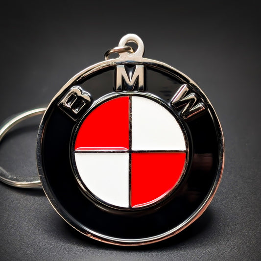 Double-sided metal BMW badge keychain with a black ring reading ‘BMW’ in silver lettering, encircling a roundel in alternating red and white quadrants, chrome split ring, on a dark surface.