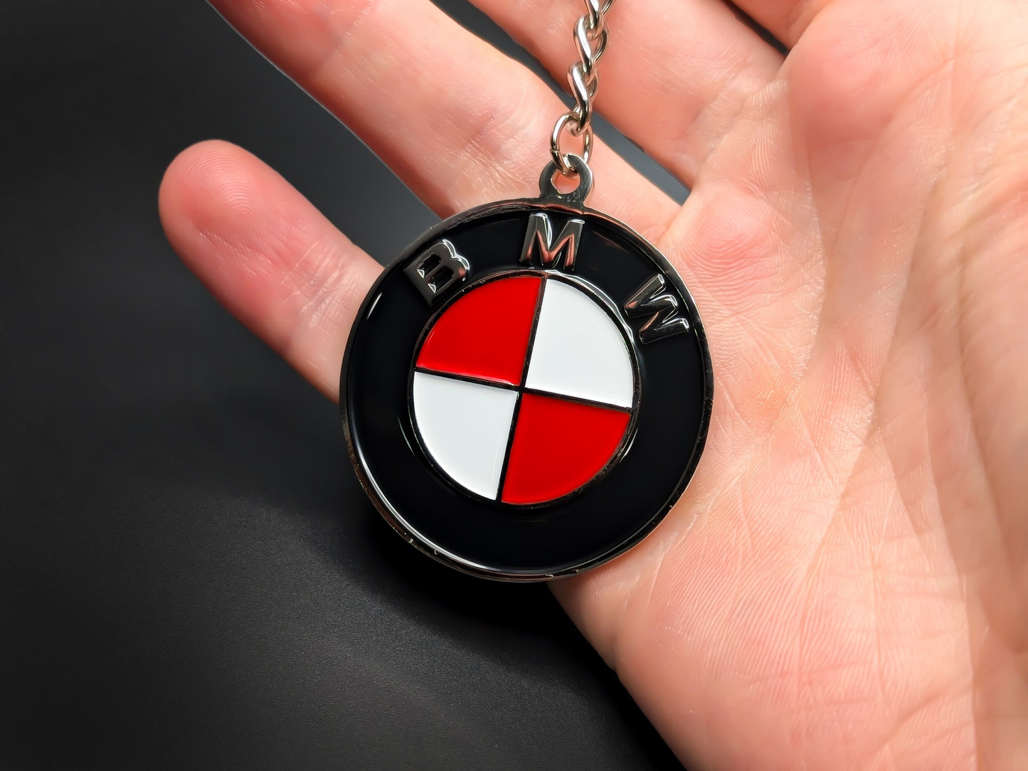 Hand holding a circular red and white BMW enamel badge keychain to demonstrate the premium metallic weight and 3D textures.