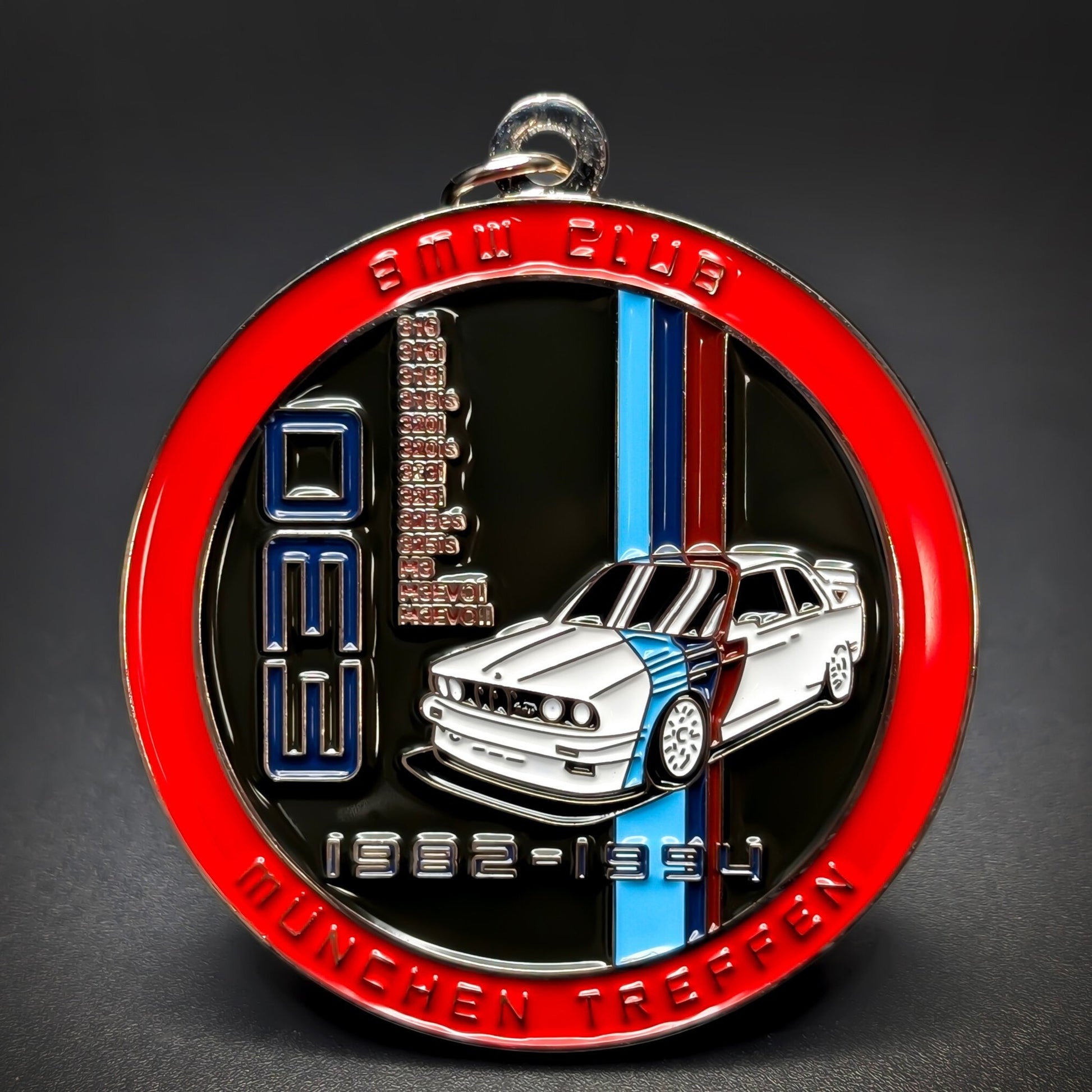 Double-sided metal BMW badge keychain with a red enamel ring reading BMW CLUB and MÜNCHEN TREFFEN, a white E30 illustration with blue, purple and red stripes on black, silver ring, on a dark surface.