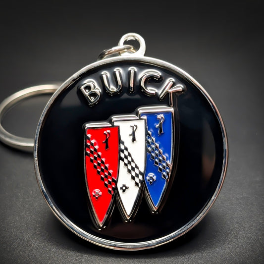 Double-sided metal Buick badge keychain, a black badge with ‘BUICK’ in chrome lettering above a red, white, and blue tri-shield emblem with checkered accents, on a split ring on a dark background.