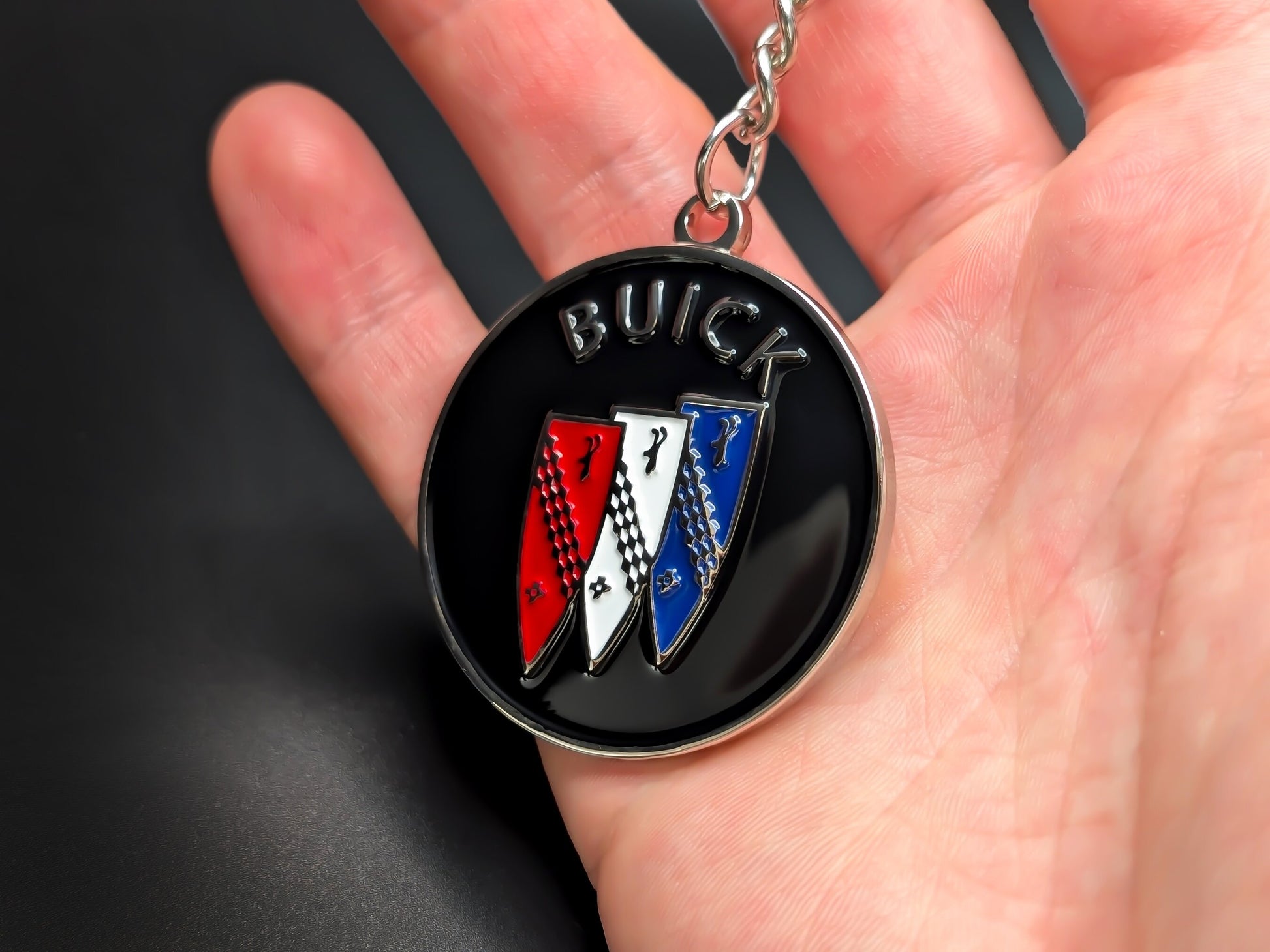 Circular Buick Tri-Shield enamel badge keychain held in hand to demonstrate the premium size, weight, and metallic shine.