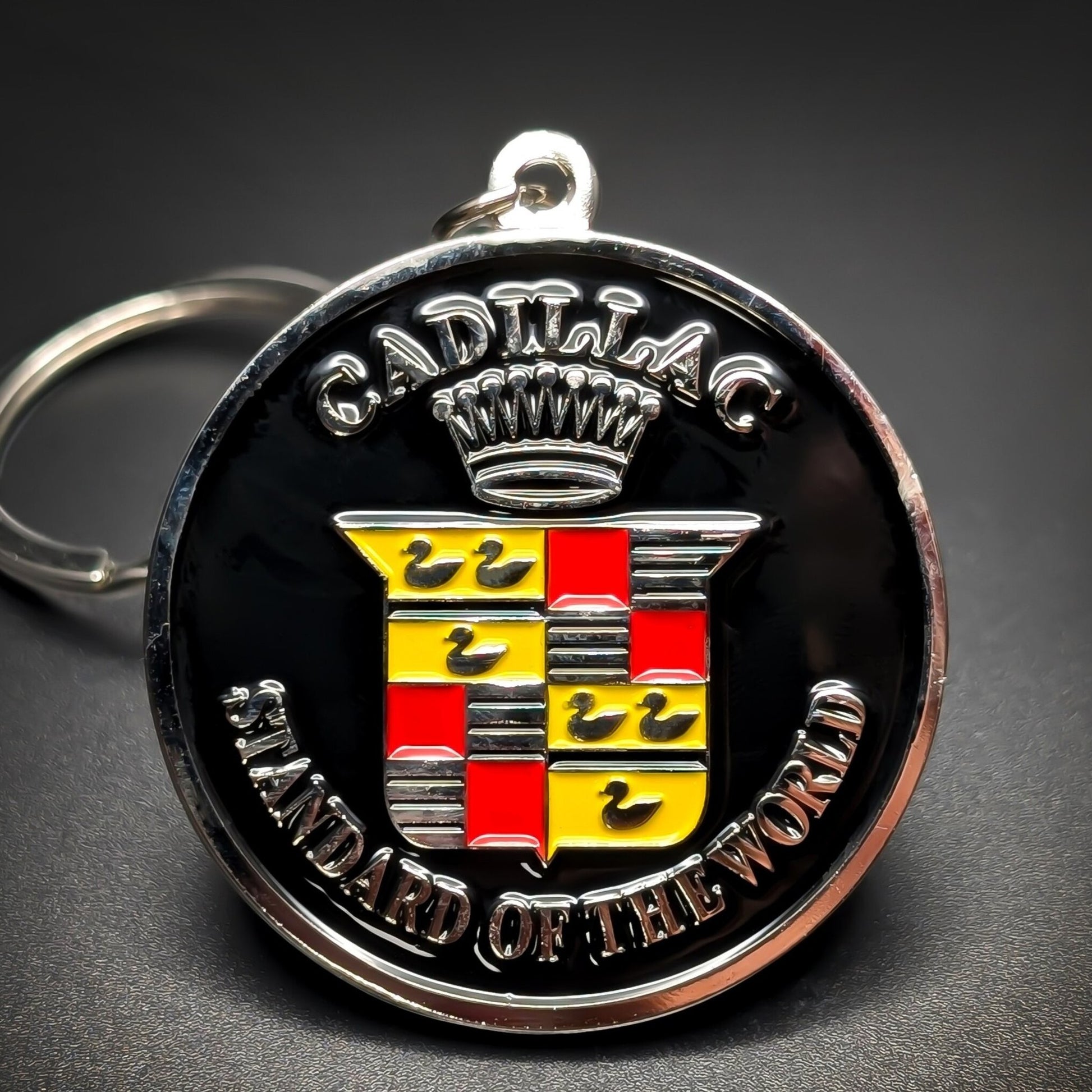 Double-sided metal Cadillac badge keychain with a black center circle, silver crown over ‘CADILLAC,’ a red-and-gold crest with black birds, ‘STANDARD OF THE WORLD’ below, split ring, dark background.