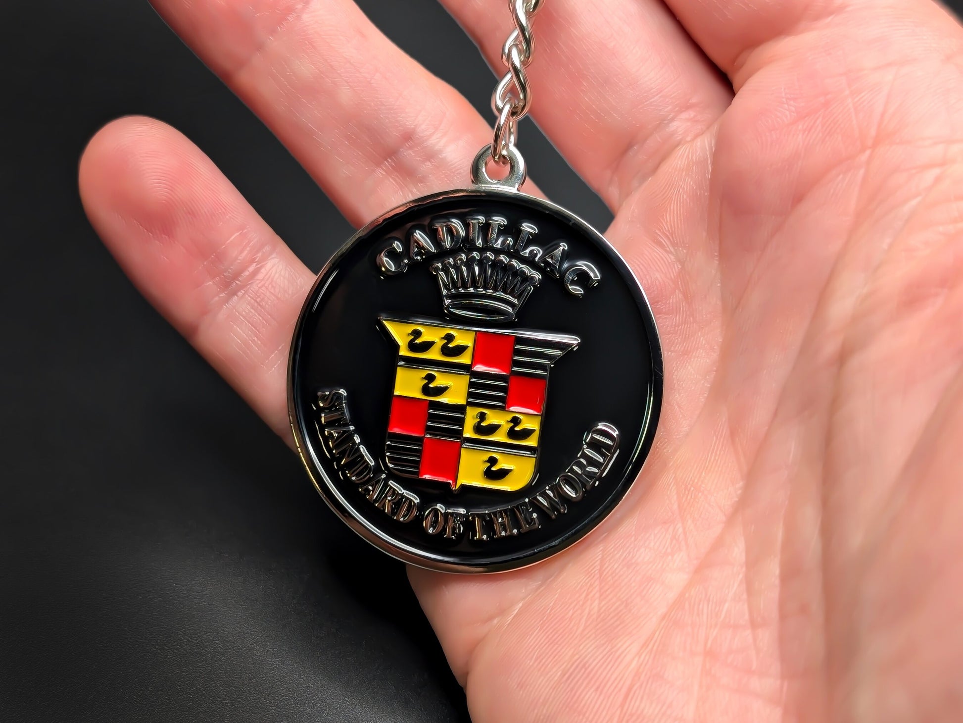 Hand holding a circular Cadillac enamel badge keychain to demonstrate the premium metallic weight and vibrant 3D raised silver details.