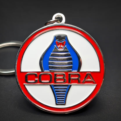 Double-sided metal Shelby Cobra badge keychain, a white medallion with a red outer ring, a chrome cobra with a blue-striped body coiled above and below COBRA in black lettering, dark surface.