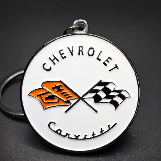 Double-sided metal Corvette badge keychain, white enamel medallion with chrome trim, “Chevrolet” arched at top and “Corvette” script below, crossed checkered and orange flags, silver split ring.