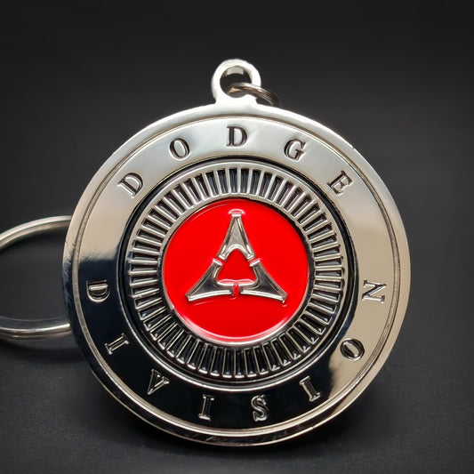 Double-sided metal Dodge Fratzog badge keychain, a chrome medallion with DODGE DIVISION lettering circling the rim and a red disc holding the silver three-pointed Fratzog emblem, dark surface.
