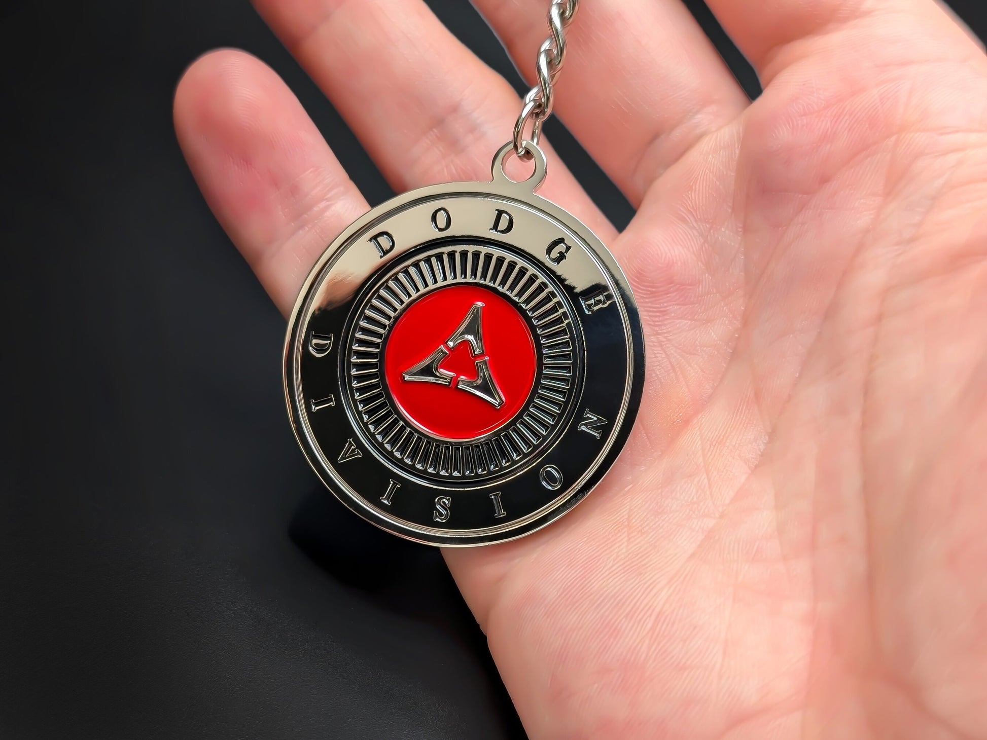 Hand holding a circular Dodge Division enamel badge keychain to demonstrate the premium metallic weight and 3D raised silver details.