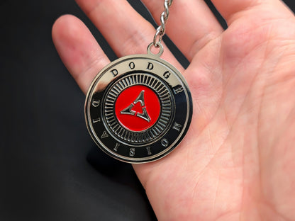 Hand holding a circular Dodge Division enamel badge keychain to demonstrate the premium metallic weight and 3D raised silver details.