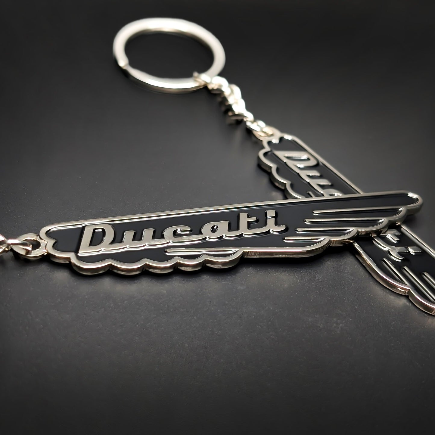 Double-sided metal Ducati badge keychain in black enamel with silver trim and cursive Ducati script, wing-shaped with a scalloped edge, attached to a split ring by a chain, on a dark surface.