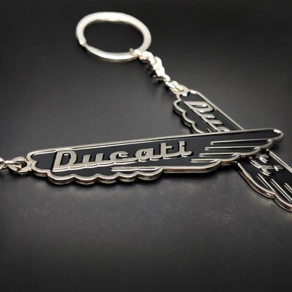 Double-sided metal Ducati badge keychain in black enamel with silver trim and cursive Ducati script, wing-shaped with a scalloped edge, attached to a split ring by a chain, on a dark surface.