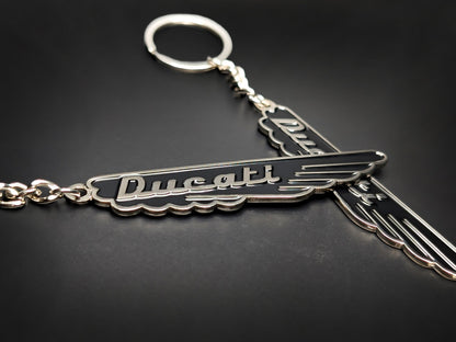 Two midnight black Ducati vintage script badge keychains with polished silver-tone wings on a dark surface.