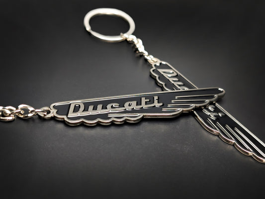Two midnight black Ducati vintage script badge keychains with polished silver-tone wings on a dark surface.