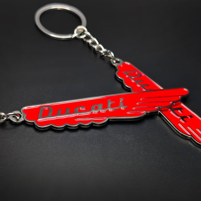 Double-sided metal Ducati badge keychain in a swept-wing shape, red enamel with silver ‘Ducati’ script and chrome trim, attached by a chain to a split ring on a dark surface.