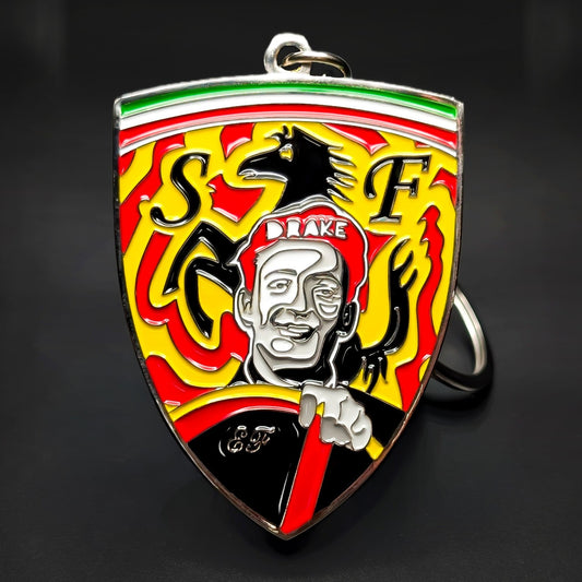 Double-sided metal Ferrari shield badge keychain, a green-white-red stripe, yellow field, black prancing horse, ‘S’ and ‘F’ initials, a portrait in a cap reading ‘DRAKE,’ split ring, dark background.