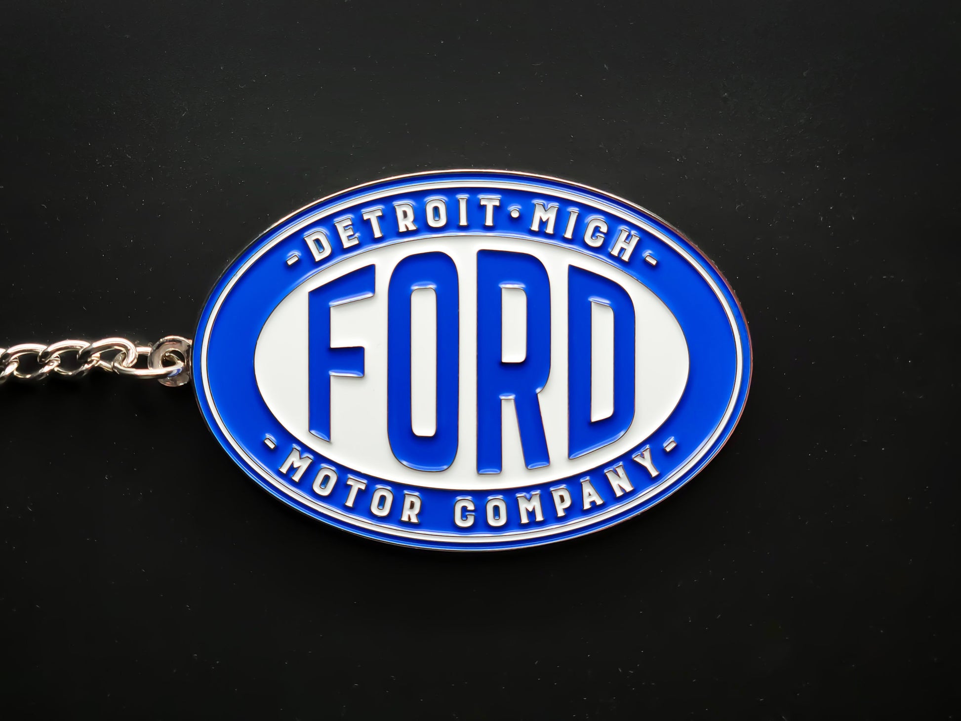 Overhead view of a Ford Detroit Motor Company keychain with a sturdy silver linked chain against a dark backdrop.