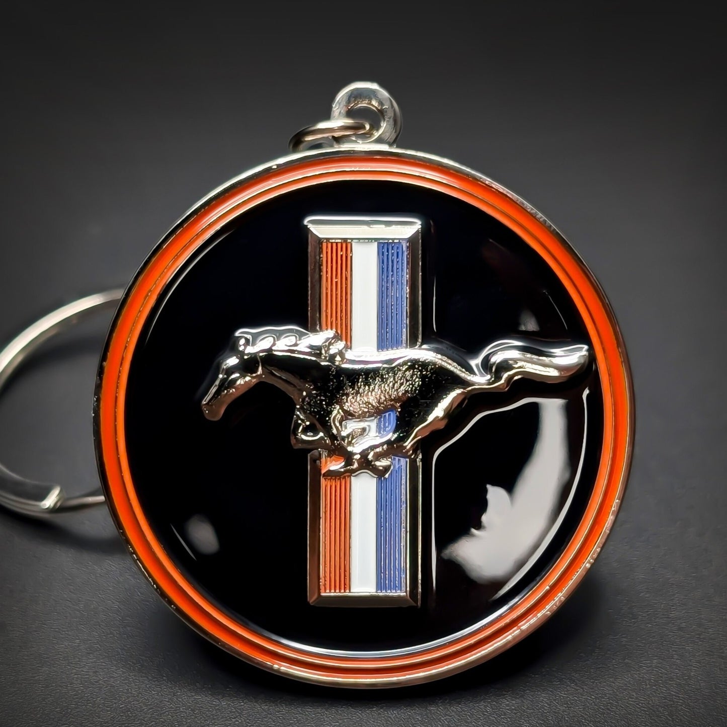 Double-sided metal Ford Mustang badge keychain, black enamel medallion with an orange and chrome rim, a red, white, and blue tri-bar, and a chrome running horse emblem at center, silver split ring.