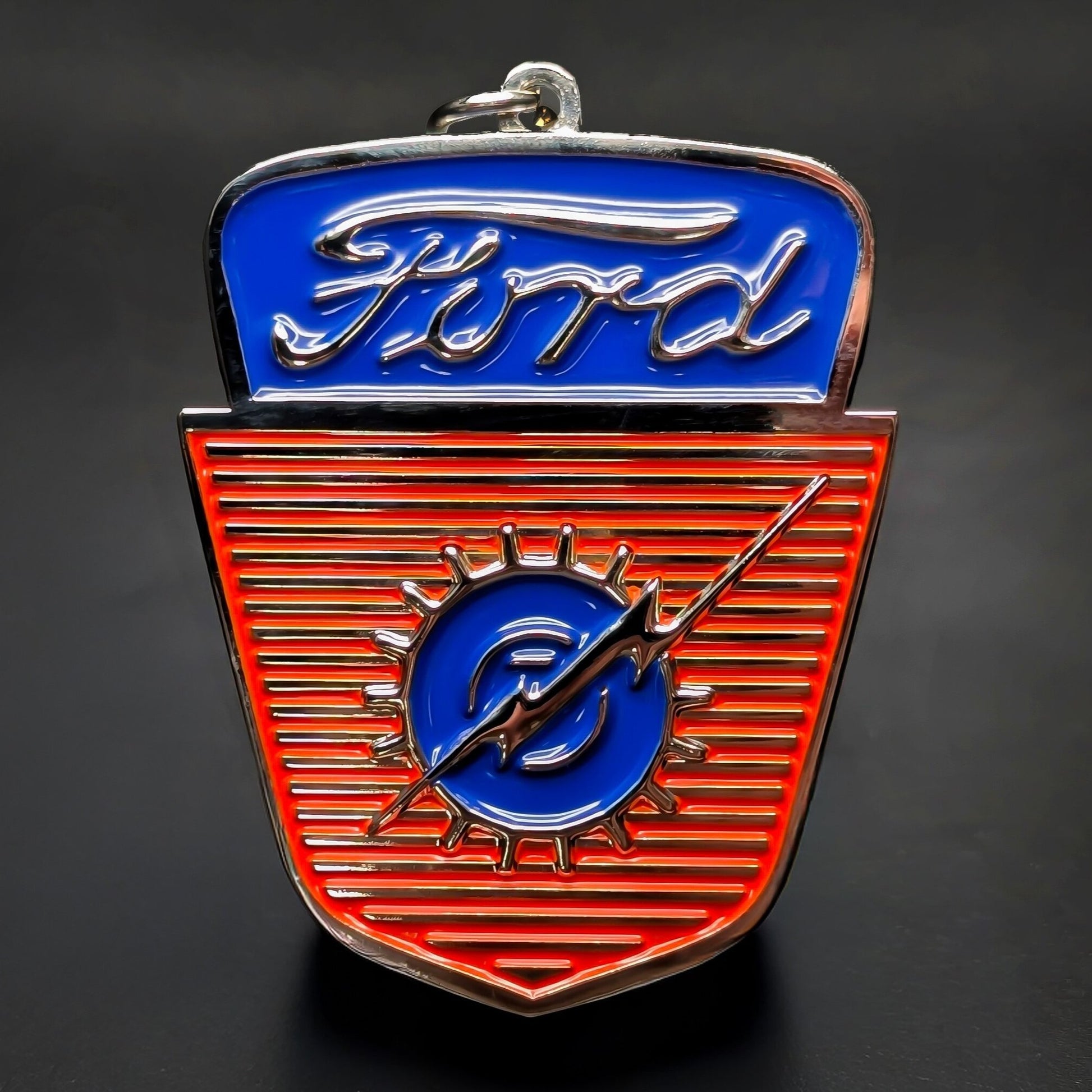 Double-sided metal Ford crest badge keychain with ‘Ford’ in silver script on blue, a red ridged shield below, blue circle, silver gear outline, lightning-bolt emblem, split ring, dark background.