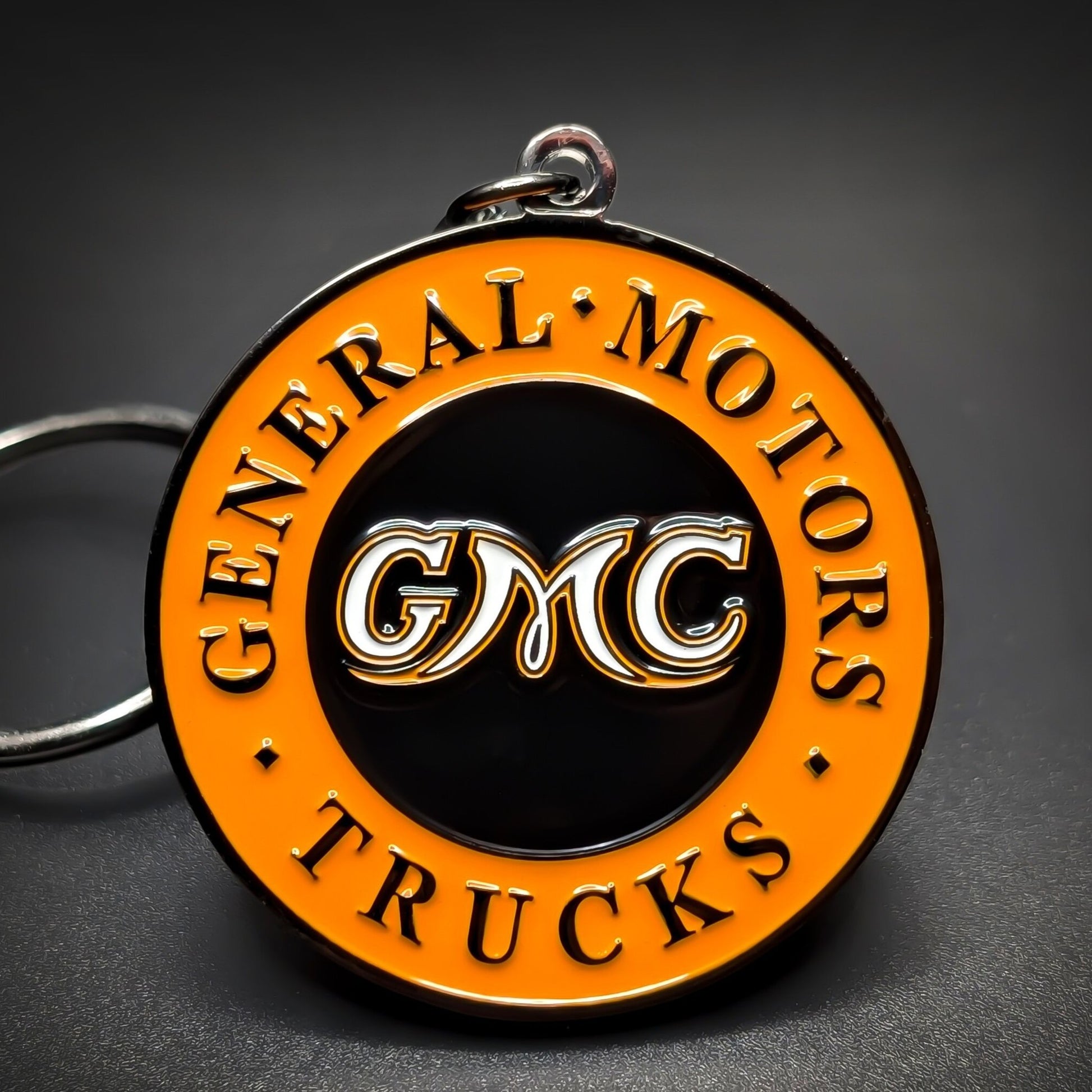 Double-sided metal GMC badge keychain, round orange and black enamel medallion with “General Motors Trucks” lettering around the rim and “GMC” in white at center, on a silver split ring.