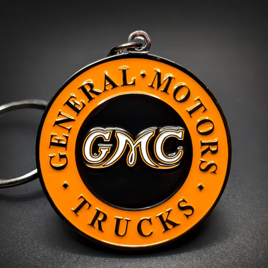 Double-sided metal GMC badge keychain, round orange and black enamel medallion with “General Motors Trucks” lettering around the rim and “GMC” in white at center, on a silver split ring.