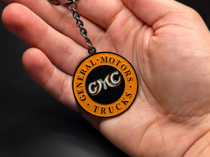 Hand holding a circular GMC Trucks enamel badge keychain to demonstrate the premium metallic weight and vibrant color finish.