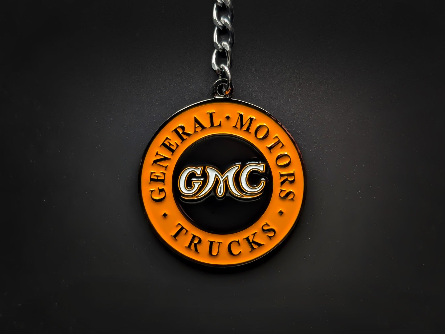 Overhead view of a GMC Trucks heritage keychain with a sturdy linked chain against a dark backdrop, highlighting the 3D raised lettering.