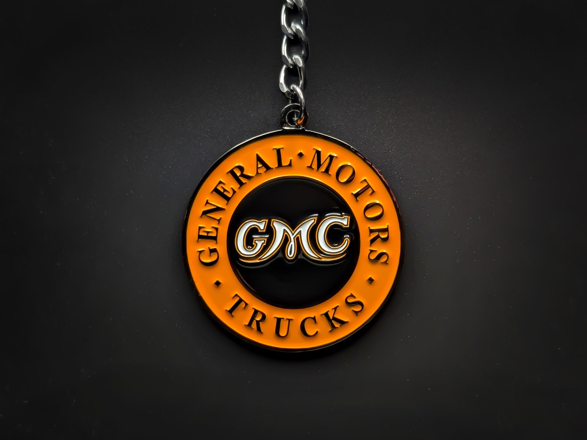 Overhead view of a GMC Trucks heritage keychain with a sturdy linked chain against a dark backdrop, highlighting the 3D raised lettering.