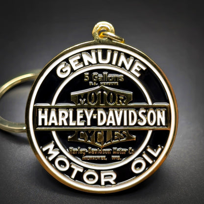 Double-sided metal Harley-Davidson badge keychain, gold-tone medallion, black enamel, white lettering GENUINE MOTOR OIL around the rim, HARLEY-DAVIDSON at center, gold split ring, dark surface.