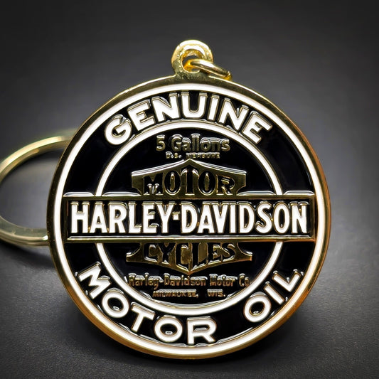 Double-sided metal Harley-Davidson badge keychain, gold-tone medallion, black enamel, white lettering GENUINE MOTOR OIL around the rim, HARLEY-DAVIDSON at center, gold split ring, dark surface.