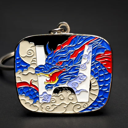 Double-sided metal Honda badge keychain with a blue dragon and red flame accents wrapped around a white H emblem on black enamel, cream cloud details, silver ring, on a dark surface.