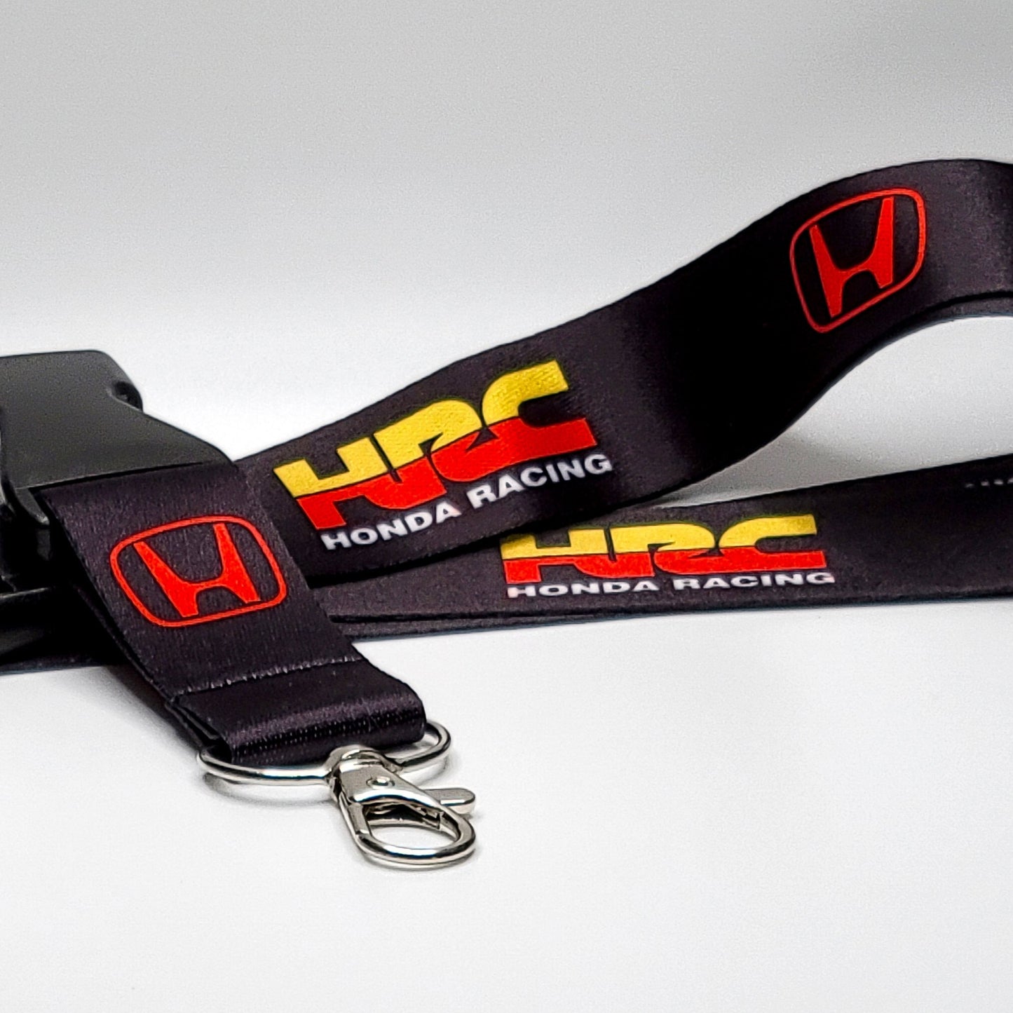 Black Honda lanyard with a red Honda H logo, a yellow-and-red HRC logo, and white Honda Racing text, coiled with a black breakaway clip and silver clasp on a light grey surface.