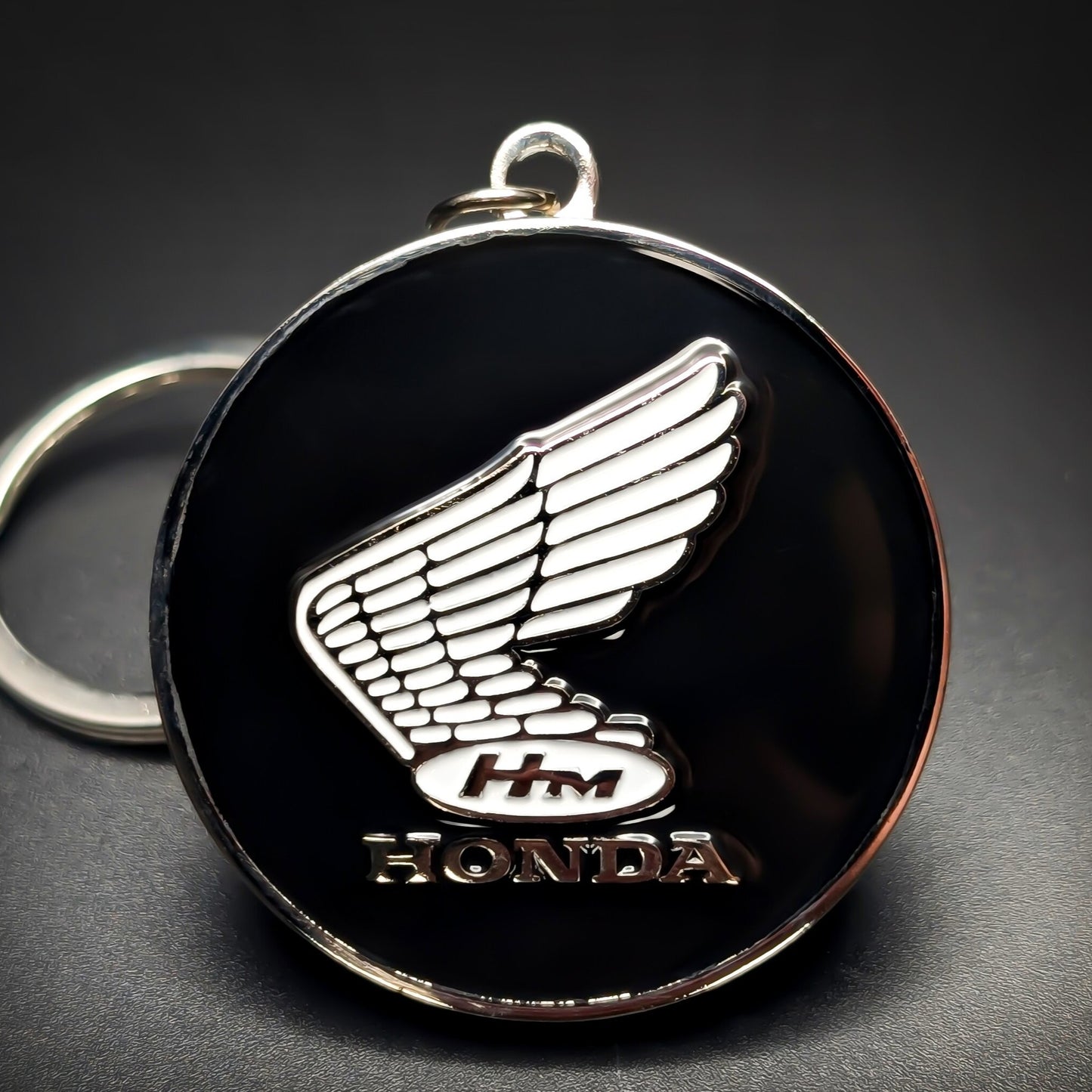 Double-sided metal Honda badge keychain with a chrome wing emblem, an ‘HM’ oval, and ‘HONDA’ lettering on glossy black enamel, chrome split ring attached, on a dark gradient background.