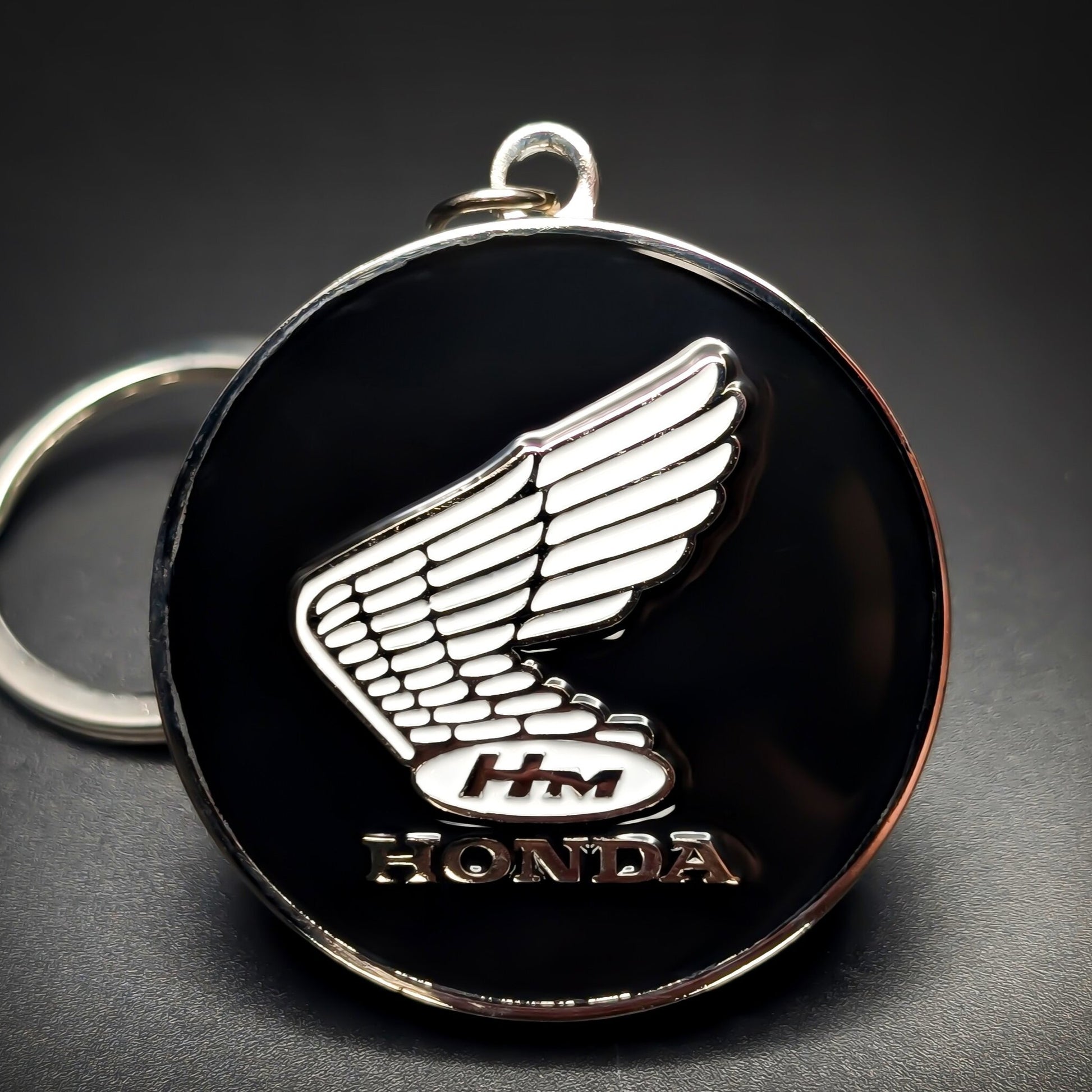 Double-sided metal Honda badge keychain with a chrome wing emblem, an ‘HM’ oval, and ‘HONDA’ lettering on glossy black enamel, chrome split ring attached, on a dark gradient background.