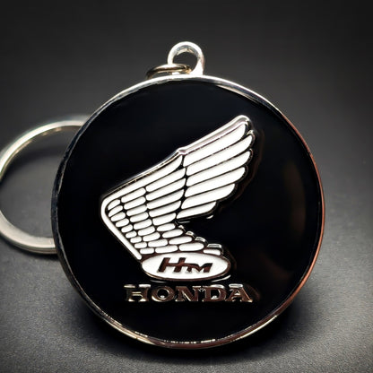 Double-sided metal Honda badge keychain with a chrome wing emblem, an ‘HM’ oval, and ‘HONDA’ lettering on glossy black enamel, chrome split ring attached, on a dark gradient background.