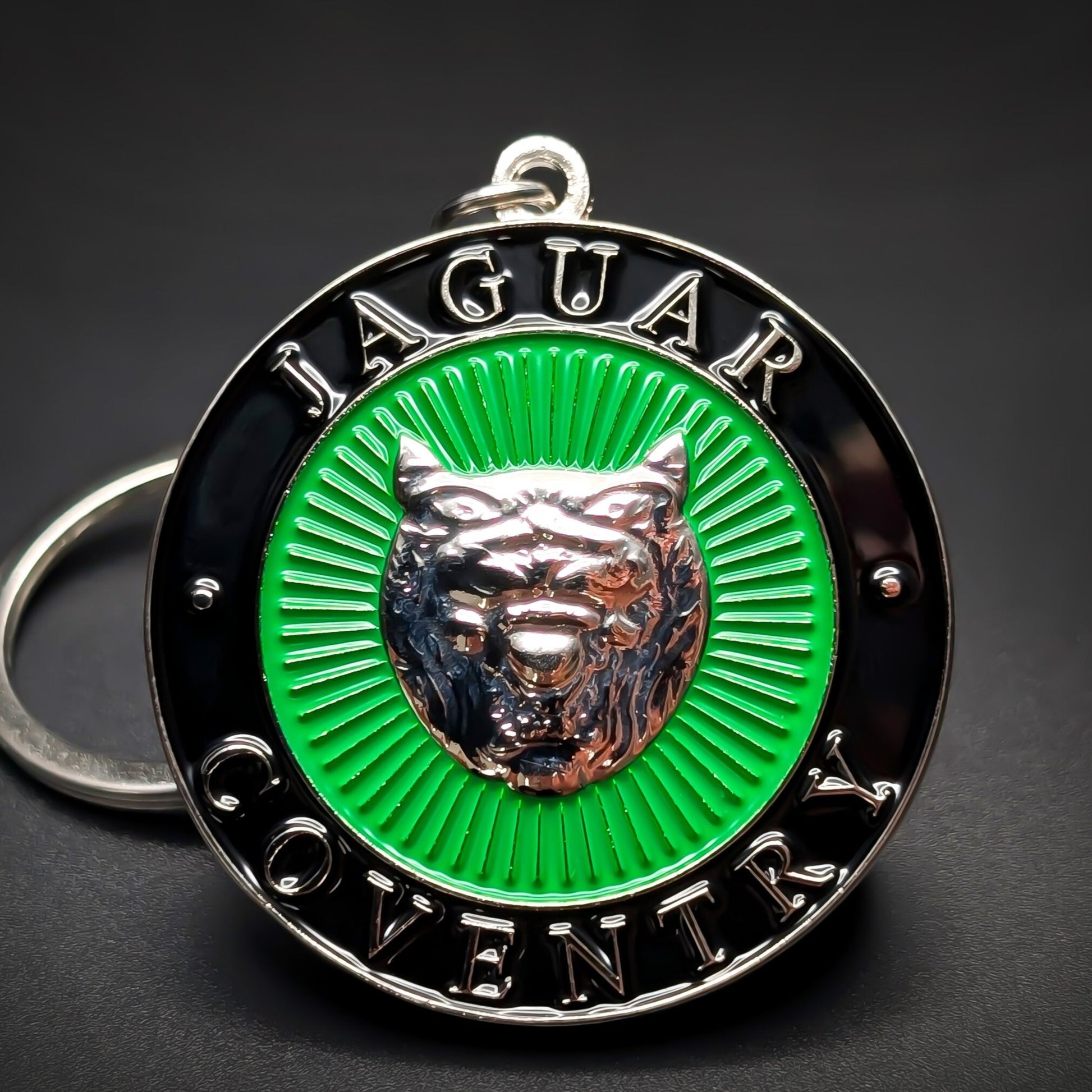 Double-sided metal Jaguar badge keychain, a black roundel with ‘JAGUAR’ and ‘COVENTRY’ lettering around a green sunburst center and silver jaguar head, on a jump ring against a dark background.