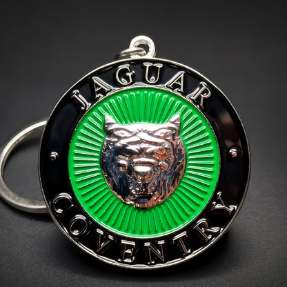 Double-sided metal Jaguar badge keychain, a black roundel with ‘JAGUAR’ and ‘COVENTRY’ lettering around a green sunburst center and silver jaguar head, on a jump ring against a dark background.