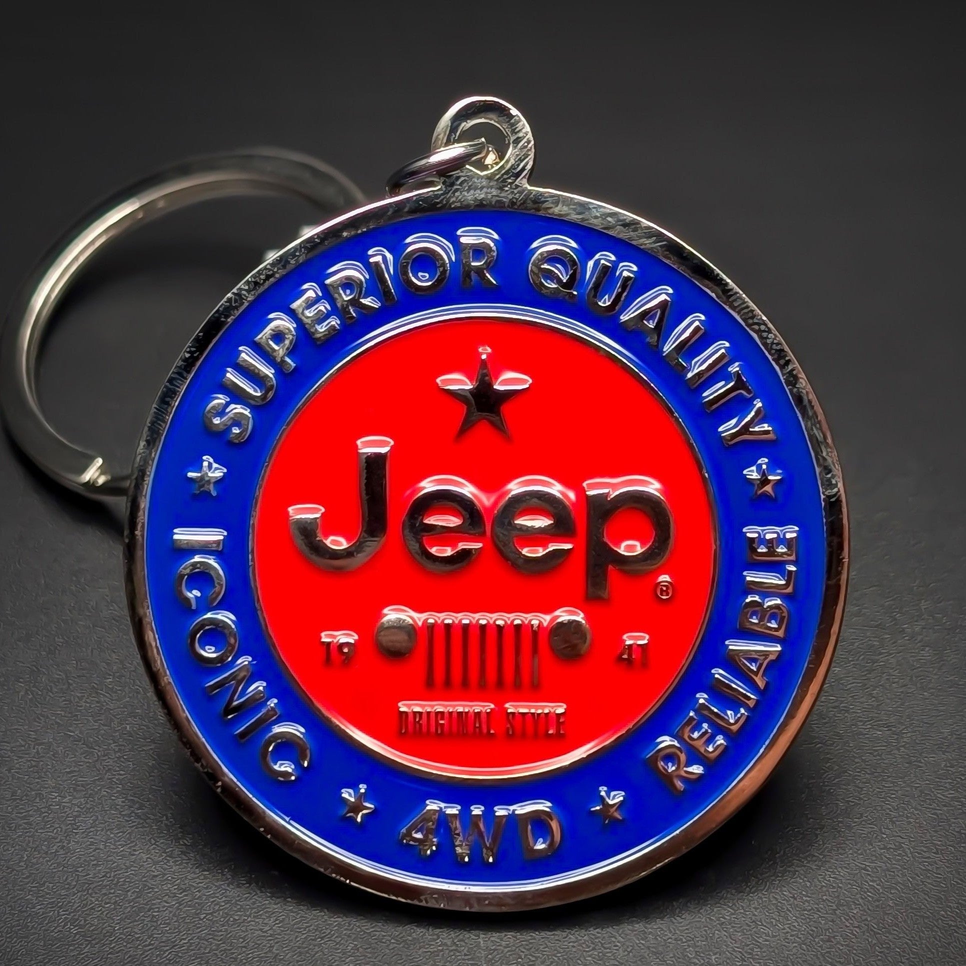 Double-sided metal Jeep badge keychain with a red center circle, black ‘Jeep’ script over a seven-bar grille icon, ringed in blue: ‘SUPERIOR QUALITY ICONIC RELIABLE 4WD,’ split ring, dark background.
