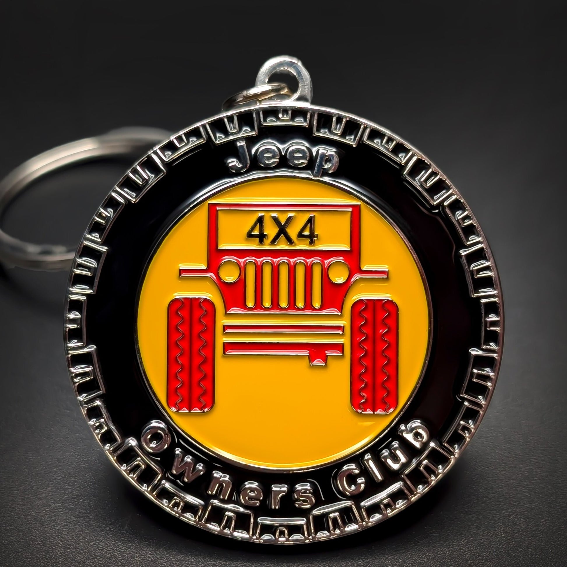 Double-sided metal Jeep badge keychain with a tire-tread silver rim reading Jeep Owners Club, a red Jeep grille illustration marked 4X4 on yellow enamel, silver split ring, on a dark surface.