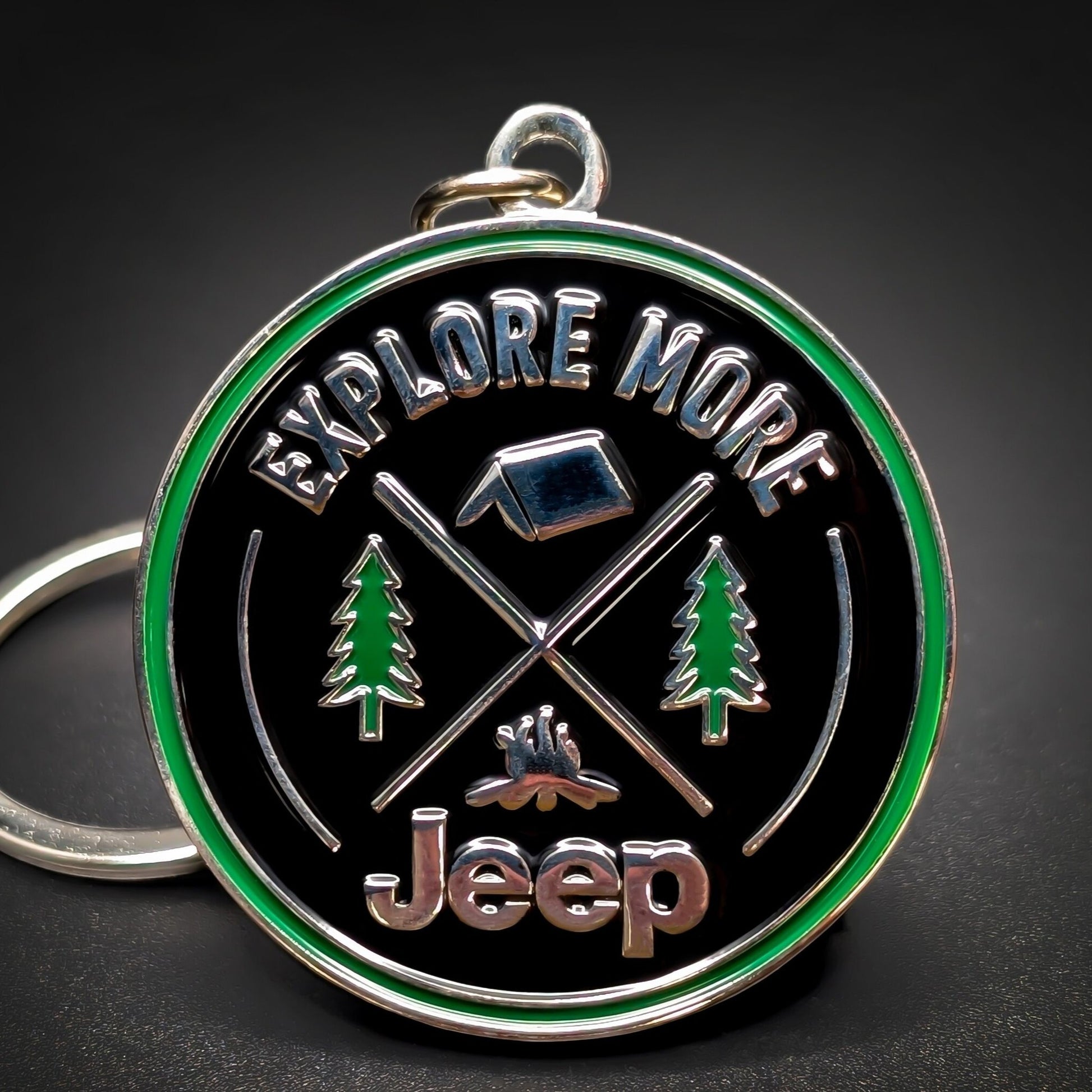 Double-sided round metal Jeep badge keychain, black enamel face and green outer ring reading EXPLORE MORE, chrome tent, crossed poles, and campfire between pine trees, chrome Jeep script below, on a hanging loop.