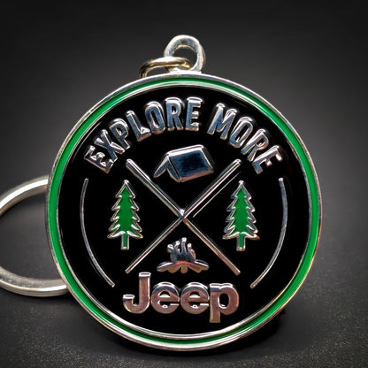 Double-sided round metal Jeep badge keychain, black enamel face and green outer ring reading EXPLORE MORE, chrome tent, crossed poles, and campfire between pine trees, chrome Jeep script below, on a hanging loop.