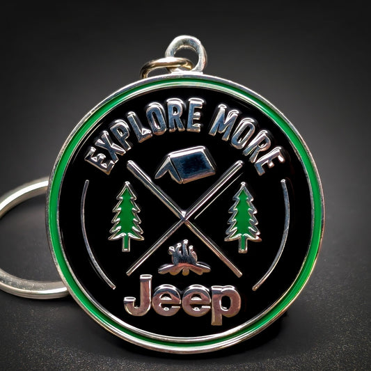 Double-sided round metal Jeep badge keychain, black enamel face and green outer ring reading EXPLORE MORE, chrome tent, crossed poles, and campfire between pine trees, chrome Jeep script below, on a hanging loop.