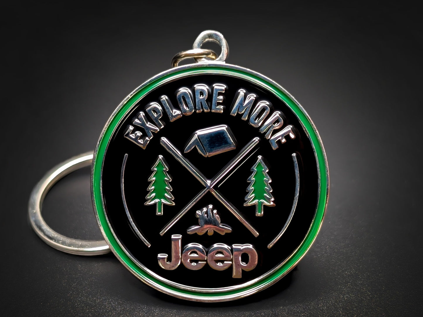 Jeep "Explore More" badge keychain featuring 3D camping icons, pine trees, and a green enamel border on a black background.