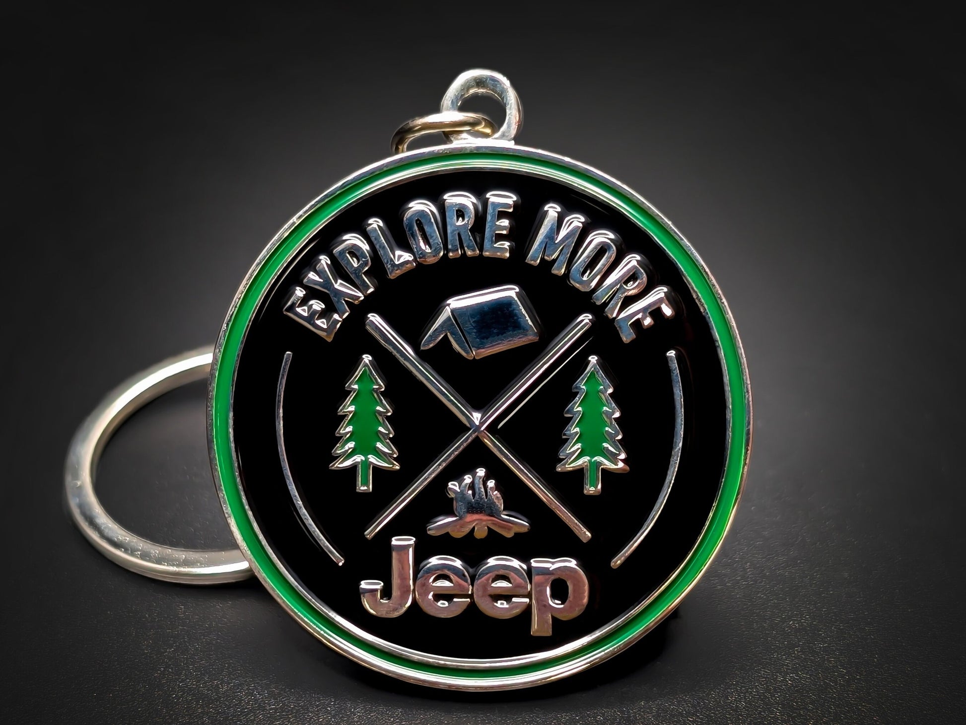 Jeep "Explore More" badge keychain featuring 3D camping icons, pine trees, and a green enamel border on a black background.